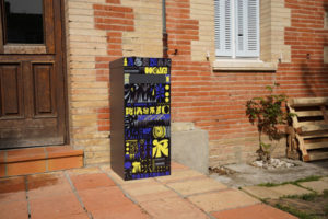 Boite à colis street art jungle avec des motifs coloré à dominante jaune bleu et noir crée par un artiste toulousain Woizo la photo a été prise devant une maison traditionnelle en brique